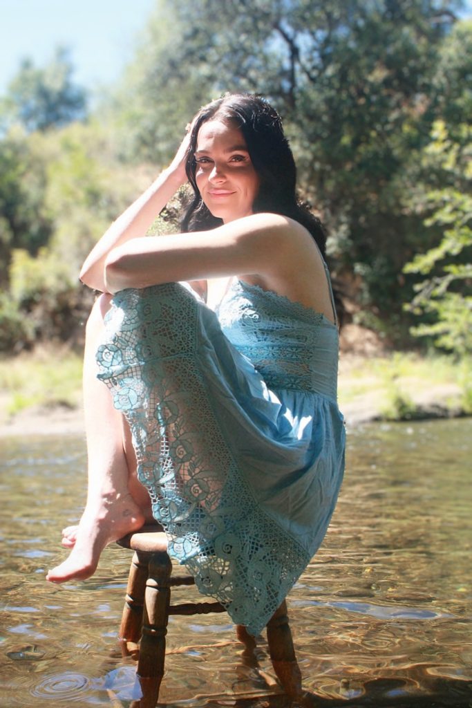 brunette woman in a blue dress posing with her legs pulled in on a stool in a river